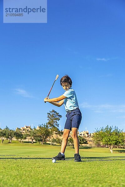 Vertikales Ganzkörperfoto eines kaukasischen Jungen  der auf einem grünen Platz gegen den blauen Himmel Golf spielt