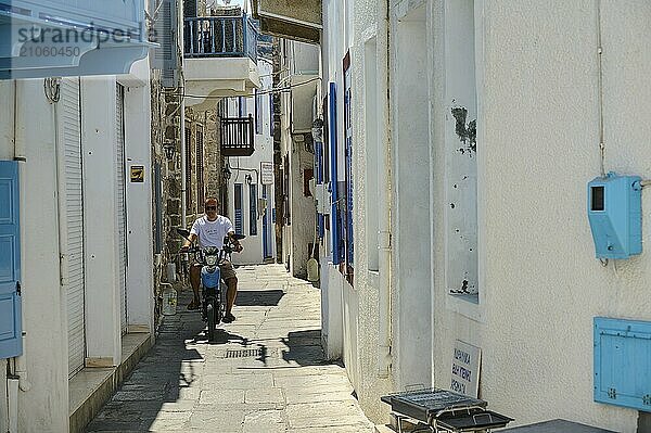 Eine schmale weiße Gasse mit blauen Fensterläden. Ein Mann fährt auf einem Motorrad durch die Sonne  Mandraki  Nisyros  Dodekanes  Griechische Inseln  Griechenland  Europa