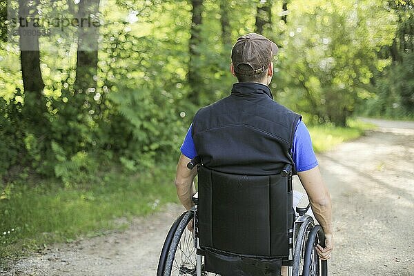 Glücklicher junger behinderter Mann in einem Rollstuhl sitzend in der Natur  der auf einem Wanderweg an einem schönen sonnigen Tag die Räder dreht