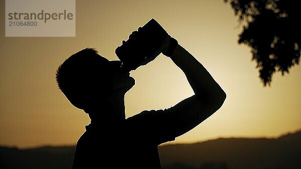 Silhouette eines fitten jungen Mannes  der Wasser trinkt  mit Sonne und Natur dahinter im Sonnenuntergang