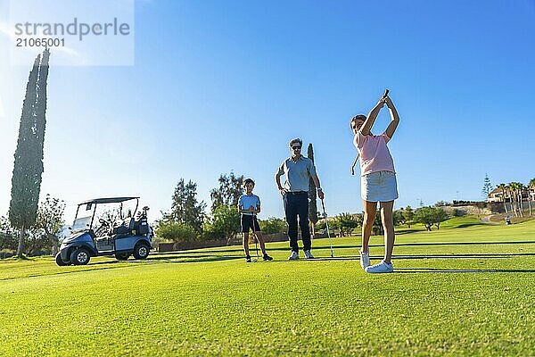 Schulmädchen schwingt Golf neben einem Jungen und einem Lehrer  der sie auf einem grünen Platz ansieht