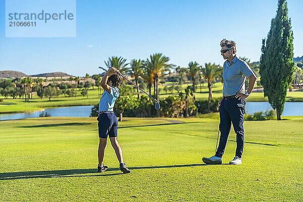 Männlicher erwachsener kaukasischer Golflehrer  der einem Jungen auf einem grünen Platz an einem sonnigen Tag gegen den blauen Himmel heißes Spiel beibringt