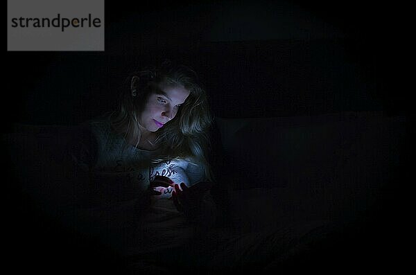 Großes Konzept der Interaktivität durch soziale Medien mit Smartphone  Frau mit Smartphone in der Nacht im Bett  Porträt in low key  schwarzer Hintergrund  wenig Licht. Farbbild