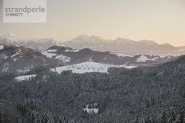 Schöne Sonnenaufgang Landschaft von Saint Thomas Kirche in Slowenien auf dem Hügel in den Morgennebel mit rosa und blauen Himmel und Triglav Berg Hintergrund