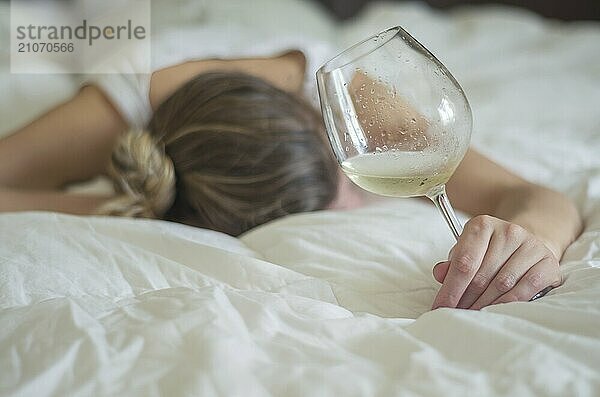 Großartiges Konzept des Alkoholmissbrauchs. Junge Frau  blondes Haar  ohnmächtig im Bett  nachdem sie zu viel Alkohol getrunken hat. Ein Glas Wein in der Hand  eine Flasche Wein