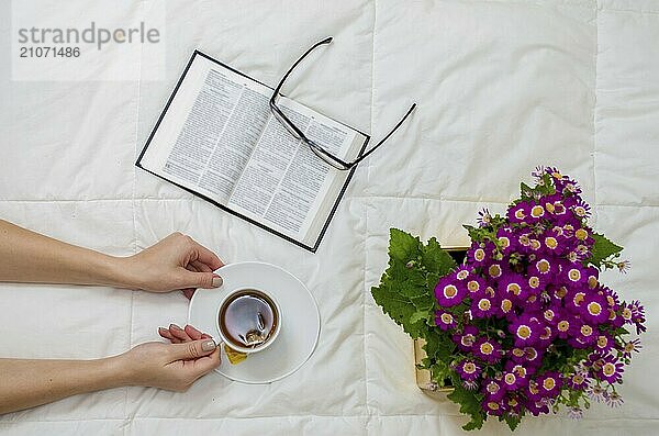 Arm einer Frau mit Tasse schwarzem Tee  Brille  Buch und lila Blumen auf weißem Bett