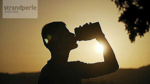 Silhouette eines fitten jungen Mannes  der Wasser trinkt  mit Sonne und Natur dahinter im Sonnenuntergang