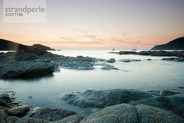 A scenic sunset along rocky Mediterranean coastline of Corsica with sailbaots on anchor in the distance; near the port town of Centuri.