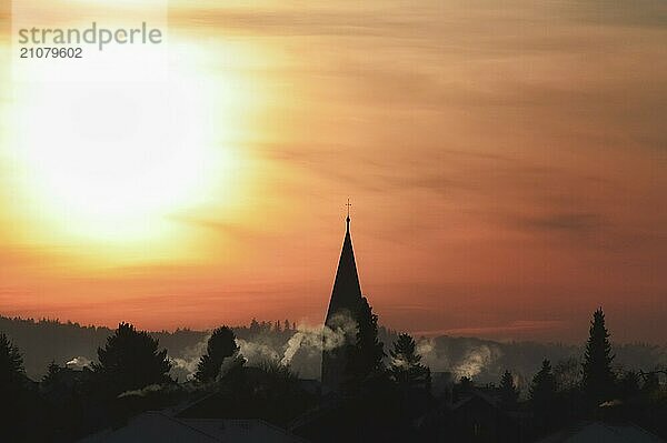 Silhouette eines Dorfes mit dem Kirchturm und Häusern mit rauchenden Schornsteinen  bei Sonnenaufgang  gegen einen goldenen Himmel  an einem Wintermorgen  in Deutschland