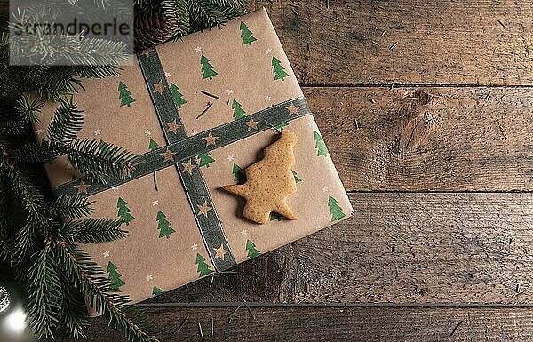 In klassisches braunes Papier eingewickelte Geschenkschachtel mit Bäumen darauf  ein Lebkuchenplätzchen in Form eines Weihnachtsbaums und Tannenzweige auf einem alten Tisch