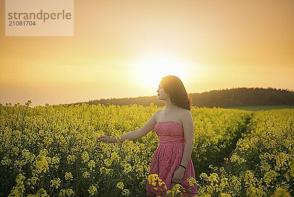 Schöne junge brünette Frau in einem rosafarbenen Kleid  die bei Sonnenuntergang durch ein blühendes Rapsfeld geht  in der Region Mähren  Tschechische Republik  Europa