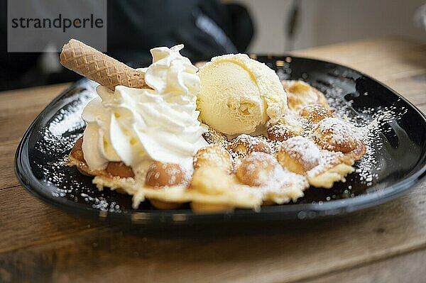 Belgische Waffel mit Vanilleeis und Sahne  Waffelröllchen  auf schwarzem Teller angerichtet und mit Puderzucker bestäubt  auf Holztisch  im Hintergrund schwarz gekleideter Mensch erkennbar  schräge Frontalsicht  Wallonien  Belgien  Europa