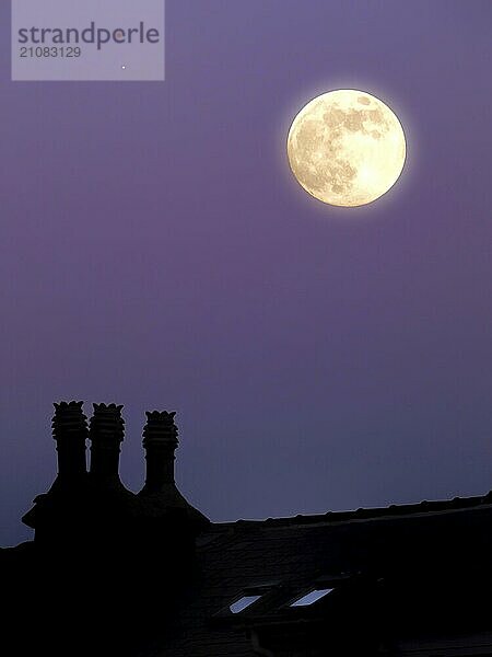 Ein glühender Vollmond in einem lila Dämmerung Sommerhimmel über einem Hausdach in der Silhouette