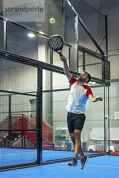 Vertikales Foto eines reifen Mannes  der springt  um den Ball beim Paddle Tennis in einer blauen Halle zu erreichen