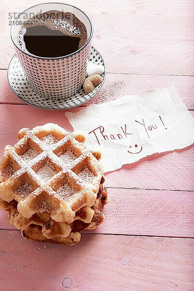 Niedliche Danksagung auf einem Stück Papier mit einem Smiley  auf einem rosa Hintergrund  neben einer heißen Tasse Kaffee und frischen Waffeln mit Puderzucker