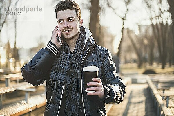 Porträt eines attraktiven Mannes mit Jacke  der ein Mobiltelefon benutzt und einen Kaffee zum Mitnehmen in der Hand hält  während er durch eine Straße in der Stadt geht