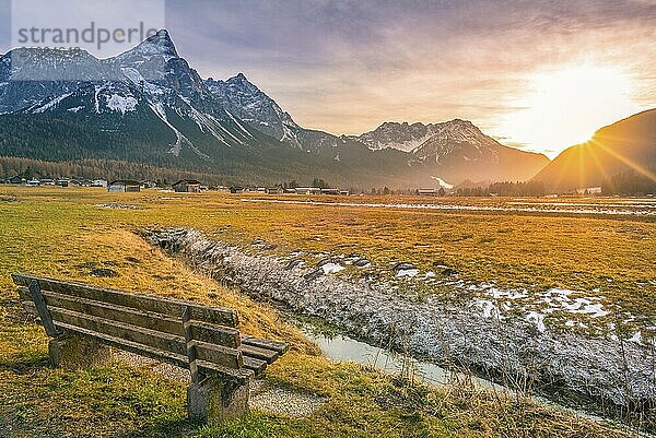 Bezaubernde Winterlandschaft mit einer Holzbank am Ufer eines kleinen Flusses mit Blick auf die österreichischen Alpen  bei Sonnenuntergang