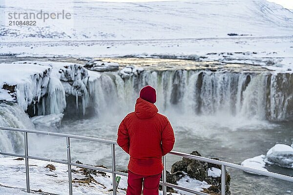 Ein Mann  der dem Godafoss Wasserfall im Winter in Island den Rücken zuwendet. Wintertourismus