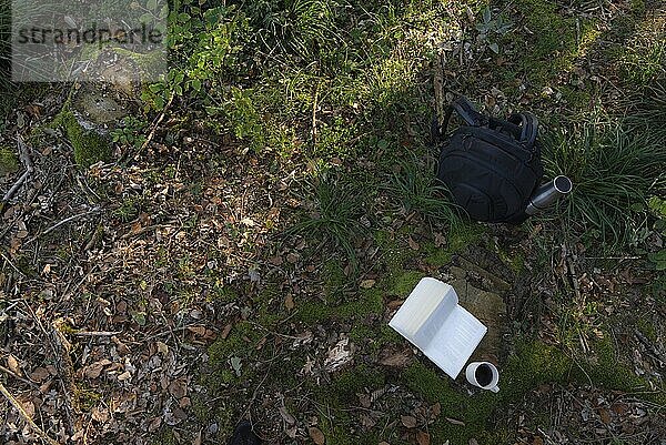 Blick von oben auf einen Wanderrucksack mit einer Thermoskanne und einem aufgeschlagenen Buch und einer Tasse Kaffee auf einer Baumwurzel  im Schatten der Bäume  in einem herbstlichen Waldboden