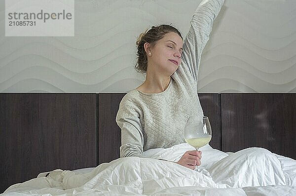 Junge blonde Frau streckt sich auf dem Bett mit einem Glas Wein in der Hand und wacht glücklich auf