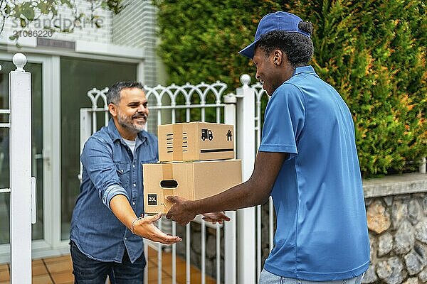 Afrikanischer Mann  der Pakete an einen reifen Mann ausliefert  der am Eingang eines Hauses in einer Wohnstraße wartet