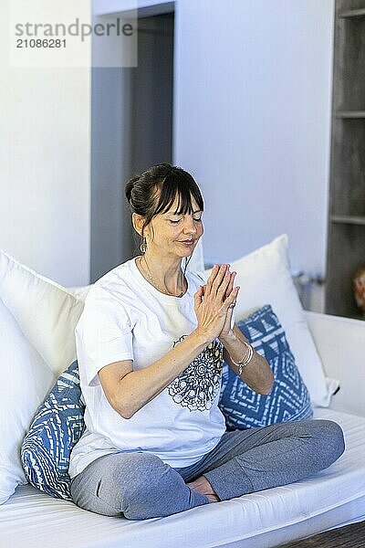 Mid-adult woman meditating on her sofa at home  with natural daylight and palms pressed together at heart center