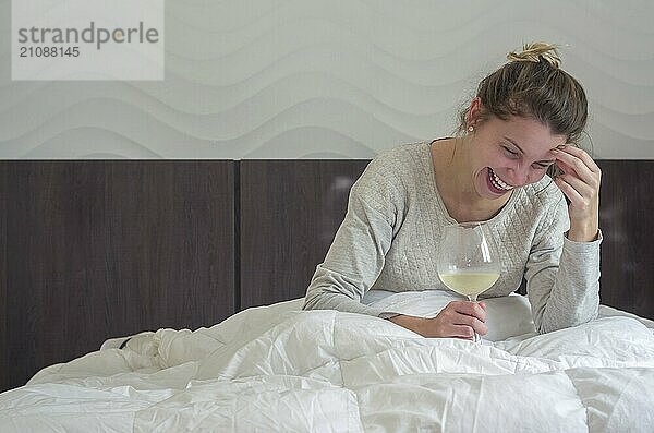 Junge blonde Frau lacht im Bett mit einem Glas Wein in der Hand und wacht glücklich auf