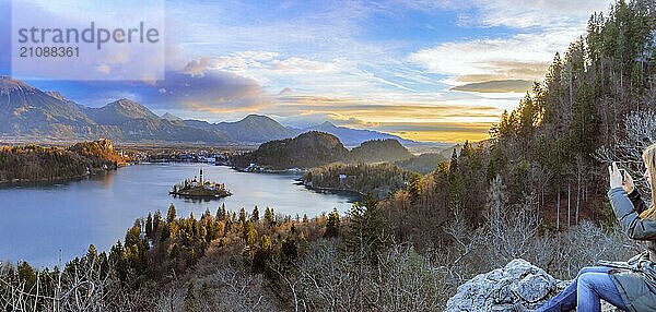 Panorama mit dem Bleder See unter einem bunten Winterhimmel bei Sonnenaufgang und einer jungen Frau  die auf einem Felsen sitzend die Aussicht genießt