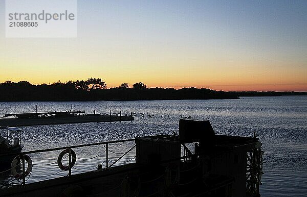 Dämmerung über einem See mit Sonnenuntergang hinter einer baumbewachsenen Insel mit leuchtend orangefarbenem Himmel und einem Steg und einem alten Paddelboot in der Silhouette