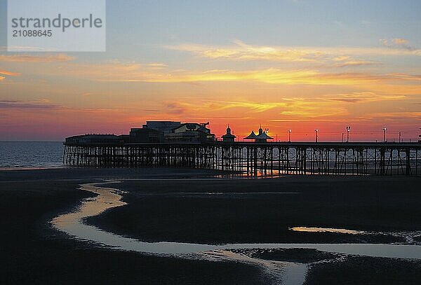 Scenic Blick auf Blackpool Nord Pier in glühenden roten Abendlicht kurz nach Sonnenuntergang mit beleuchteten rosa und gelben Himmel und Wolken mit Farben auf dem dunklen Strand in Struktur in der Silhouette reflektiert