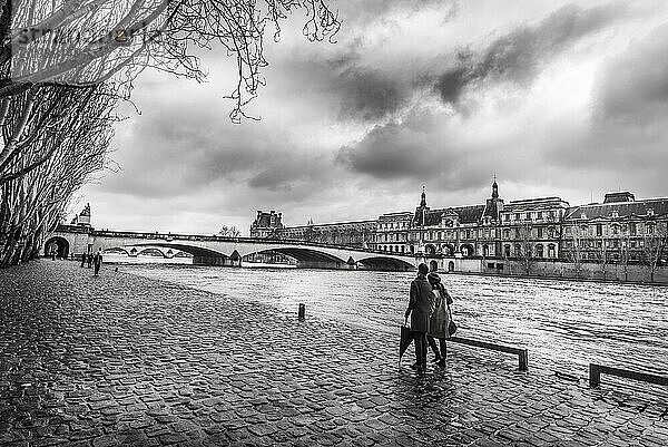 Schwarz Weiß Bild mit der Seine  ihrem Ufer  wo Menschen spazieren gehen  einer alten Brücke und dem Orsay Museum  unter einem dramatischen Himmel  an einem kühlen Februartag