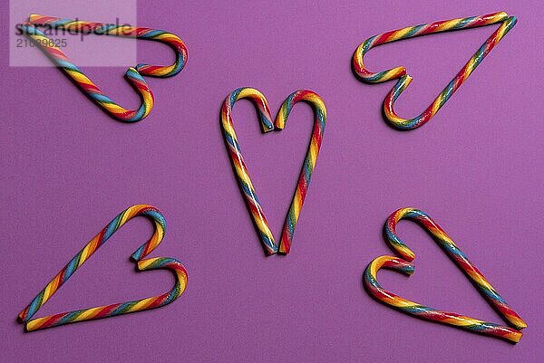 Above view of rainbow colored candy canes arranged in heart shape on a purple paper background. Minimalist Christmas concept