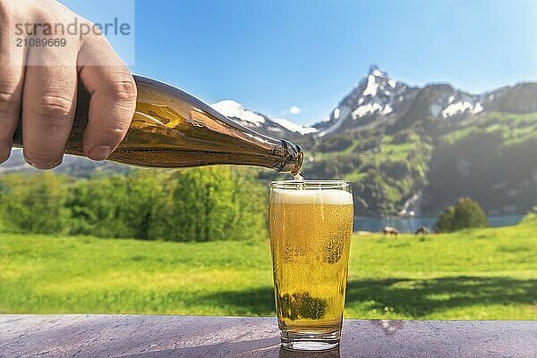 Hand eines Mannes  der Bier aus einer Flasche in ein Glas gießt  mit einer Sommerlandschaft im Hintergrund mit Bergen und grünen Wiesen  an einem sonnigen Tag