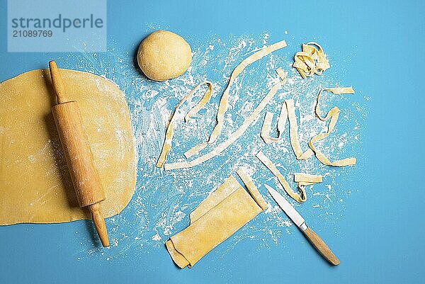 Italienische Nudelherstellung zu Hause  von Hand  auf blauem Hintergrund. Ausrollen und Schneiden von rohem Teig für die Formung von Tagliatelle. Zubereitung von langen Bandnudeln