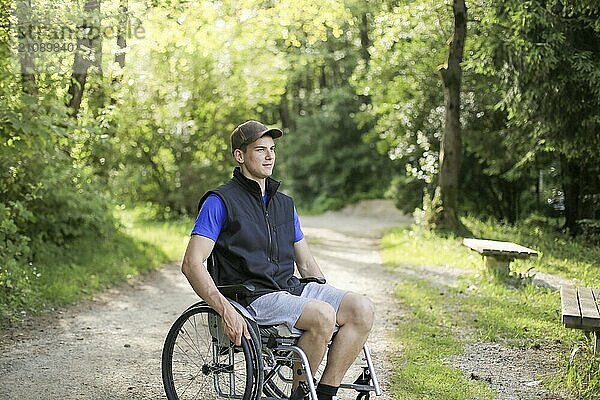 Happy and young disabled or handicapped man sitting on a wheelchair in nature turning wheels on a walking road at a beautiful sunny day