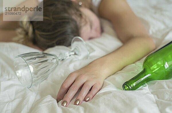 Großartiges Konzept des Alkoholmissbrauchs. Junge Frau  blondes Haar  ohnmächtig im Bett  nachdem sie zu viel Alkohol getrunken hat. Ein Glas Wein in der Hand  eine Flasche Wein