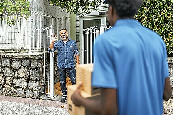 Älterer kaukasischer Mann winkt einem jungen afrikanischen Zusteller zu  der ein Paket trägt