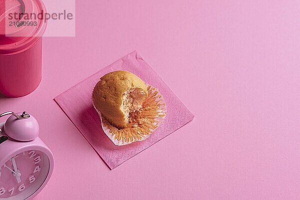 Frühstückstisch mit einem Muffin  einer Tasse Kaffee und einem Wecker. Gebissener Muffin auf rosa Hintergrund. Rosa Tasse Kaffee und Uhr