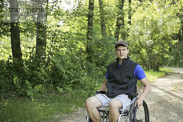 Happy and young disabled or handicapped man sitting on a wheelchair in nature turning wheels on a walking road at a beautiful sunny day