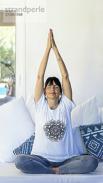 Mid-adult woman practicing meditation on her sofa at home  surrounded by natural daylight with her palms pressed together above her head