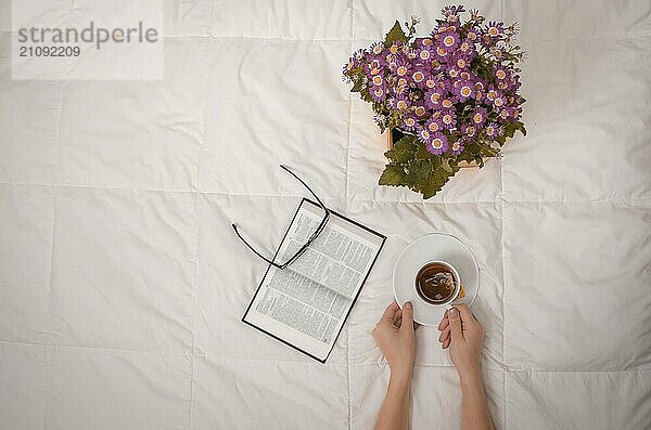 Arm einer Frau mit Tasse schwarzem Tee  Brille  Buch und lila Blumen auf weißem Bett