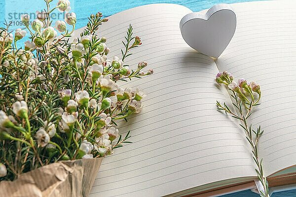 Weiße kleine Blumen und ein Holzherz auf leeren Notizbuchseiten  auf blauem Hintergrund. Nahaufnahme. Rahmen für den Valentinstag. Hochzeitsplanung Kontext