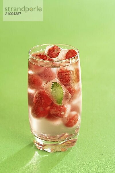 Wasser und gefrorene Himbeeren in einem Glas auf grünem Hintergrund. Gefrorene Früchte und Minzblätter in einem Glas mit Wasser