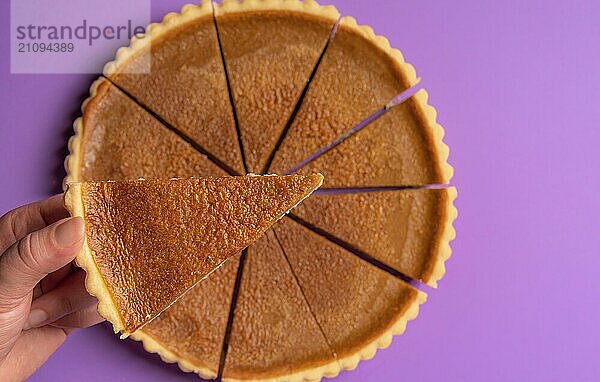 Frau hält ein Stück Kürbiskuchen  über den ganzen Kuchen  auf einem lila Hintergrund. Minimalistisches Bild. Flach gelegt. Hausgemachter Kuchen. Urlaubsdessert