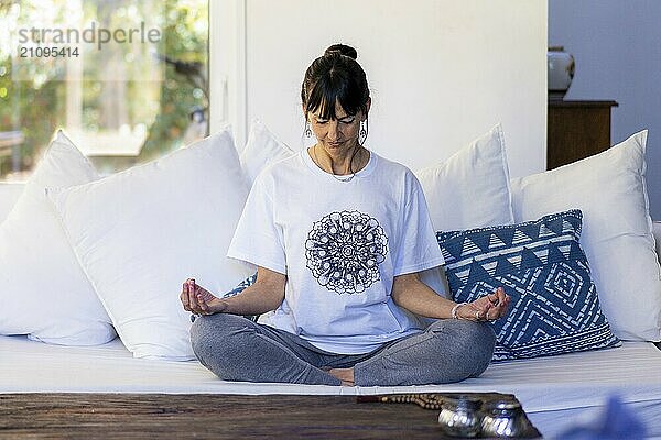 Mid-adult woman meditating in her living room  sitting on a sofa with natural daylight  her hair in bangs and tied back  hands in Gyan Mudra