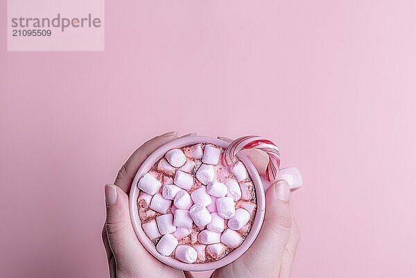 Weihnachtsgetränk Bild  eine Tasse heiße Schokolade mit rosa Mini Marshmallows und eine Xmas Zuckerstange  mit zwei Händen gehalten  über einem rosa Hintergrund