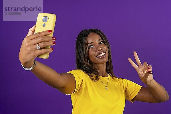 Studioporträt mit lila Hintergrund einer afrikanischen Frau  die lächelnd den Sieg erringt  während sie ein Selfie mit einem Mobiltelefon macht