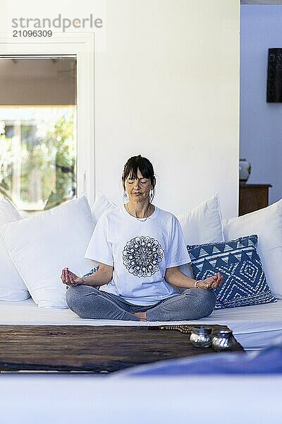 Mid-adult woman practicing meditation on her sofa during the day  illuminated by natural light  with bangs and her hair tied up  hands in Gyan Mudra