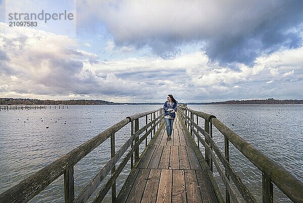 Schöne junge Frau lächelnd und zu Fuß allein auf einem langen Holzsteg  über den Chiemsee  genießen die frische Luft und den Sonnenuntergang  in Deutschland