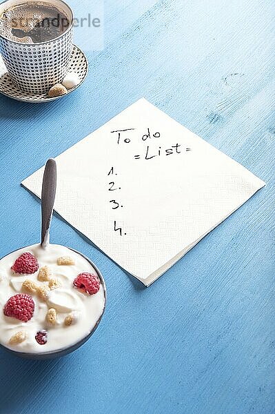 Weiße Serviette mit einem Entwurf einer To Do Liste  umgeben von einer Schale Joghurt mit Müsli und Himbeeren und einem Becher mit aromatischem Kaffee  auf blauem Hintergrund